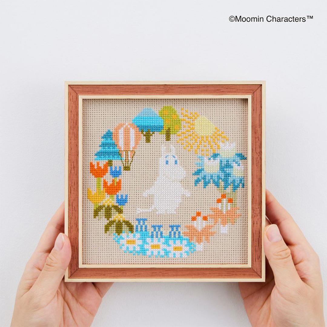 A person's hands holding a framed cross-stitch embroidery depicting a whimsical scene with a white animal, flowers, and various other colorful elements.