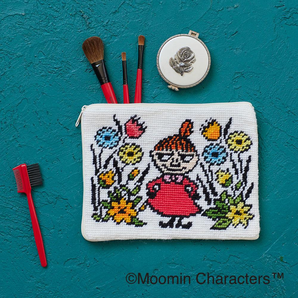 A white makeup bag with a colorful embroidered design depicting a young girl surrounded by flowers, along with a red brush and a small white compact.