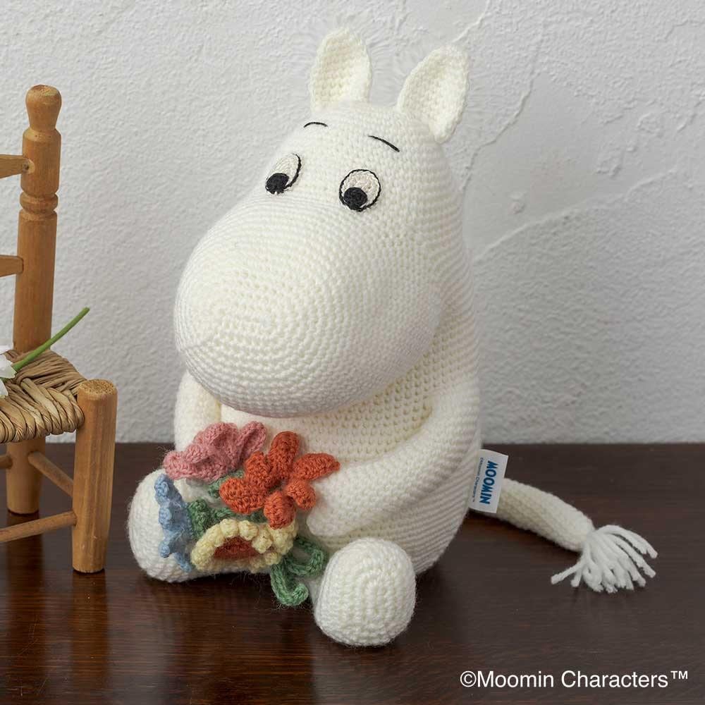 A white crocheted toy resembling a character from the Moomin characters sits on a wooden surface, holding a bouquet of flowers.