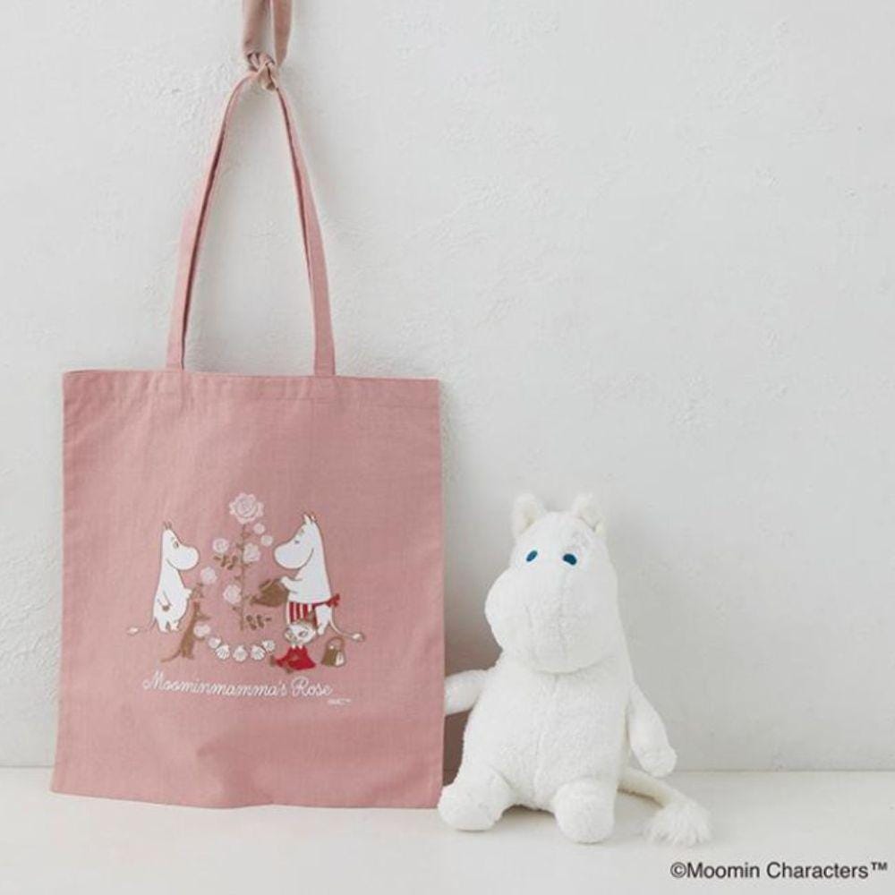 A pink tote bag with a printed design of Moomom characters and a white stuffed animal are displayed against a white wall.