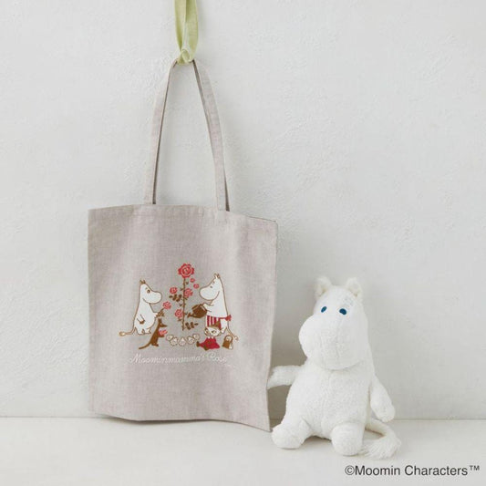 A beige tote bag with a printed design of two anthropomorphic animals, one with a flower, and the text "Moomin moomin's rose" is hanging on a wall next to a white stuffed animal.