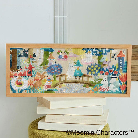 A framed cross-stitch embroidery depicting a whimsical forest scene with various animals, plants, and a bridge.