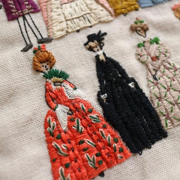 a close-up view of a hand-embroidered scene with three distinct figures, each wearing a different colored dress. The scene appears to be a part of a larger embroidered design, as suggested by the presence of other figures and patterns in the background.