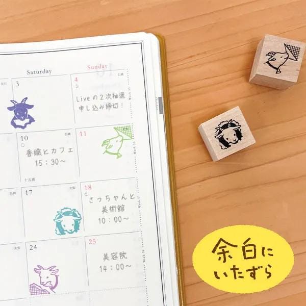 a calendar with a yellow border, a yellow sticker with Japanese text, and three wooden blocks with animal faces on a wooden surface.