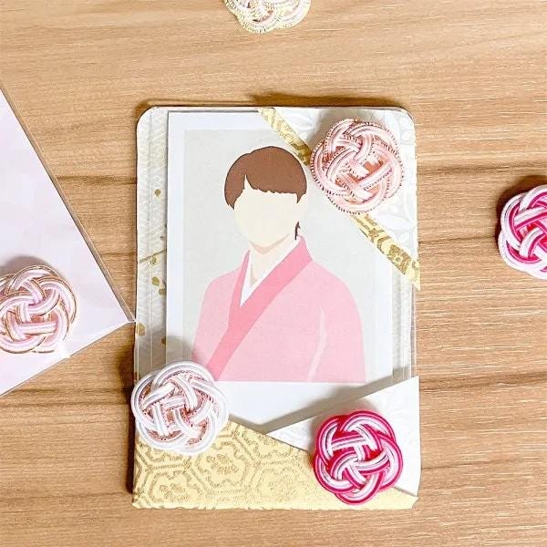 a framed illustration of a person wearing a pink kimono, surrounded by pink and white knotted ornaments on a wooden surface.