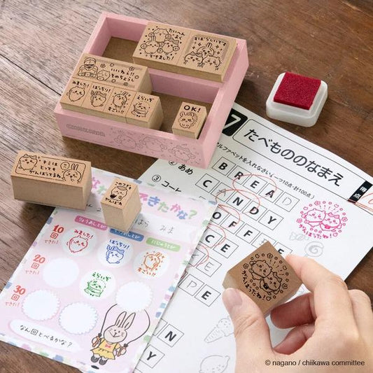 a collection of various items, including a pink stamp holder, a red stamp, and a sheet of paper with a crossword puzzle. A hand is holding a stamp in the foreground, and there are several other stamps and a stamp book visible in the background.