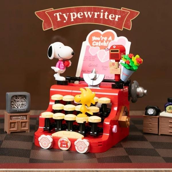 A red toy typewriter with a cartoon character, likely a Peanuts character, sitting on top of it. The typewriter has a small television and a toy mailbox on it, and there are other small toy items around the typewriter.