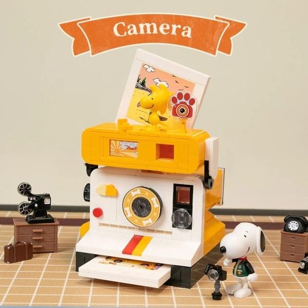 A Peanuts-themed toy set featuring a camera, a typewriter, and a Snoopy figurine, with the word "Camera" written above the set.