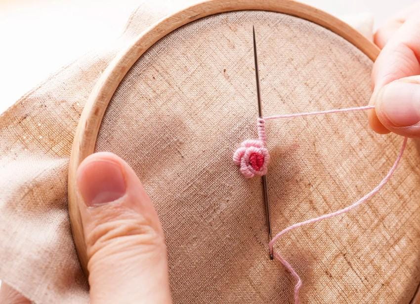 A person's hand is carefully stitching a delicate pink flower onto a piece of fabric using a needle and thread.