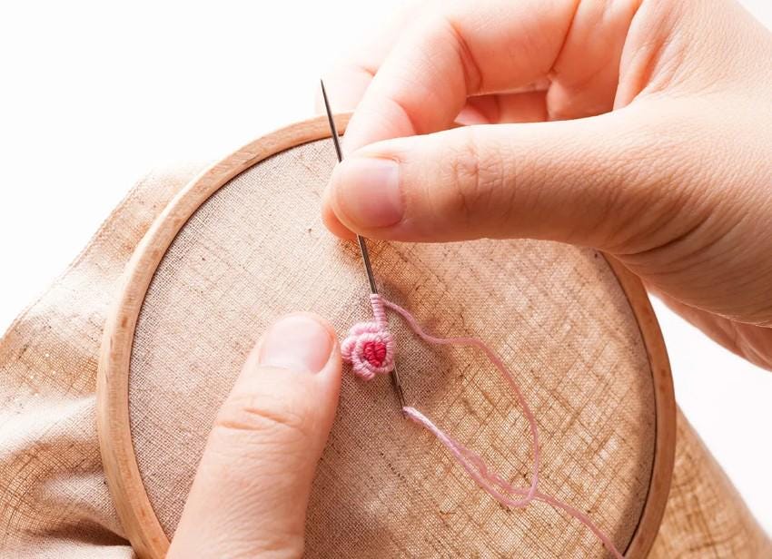 A person's hands are shown carefully stitching a small pink flower onto a piece of fabric using a needle and thread.