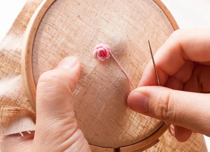 A person's hands are carefully stitching a delicate pink flower onto a piece of fabric using a needle and thread.