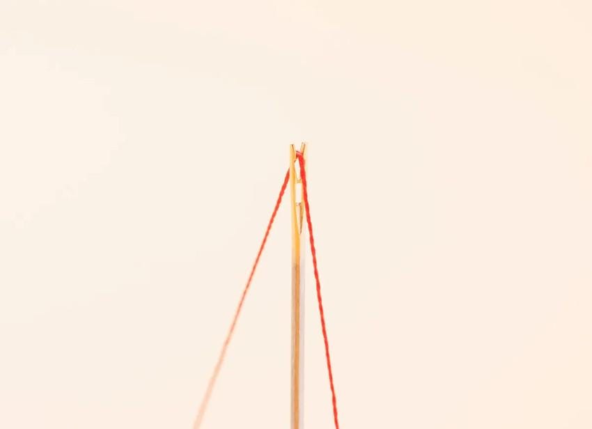 A close-up view of a needle with red thread, standing upright against a plain white background.