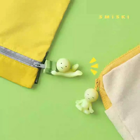 two small, glowing figurines on a green background, with a yellow zipper and a yellow object in the background.