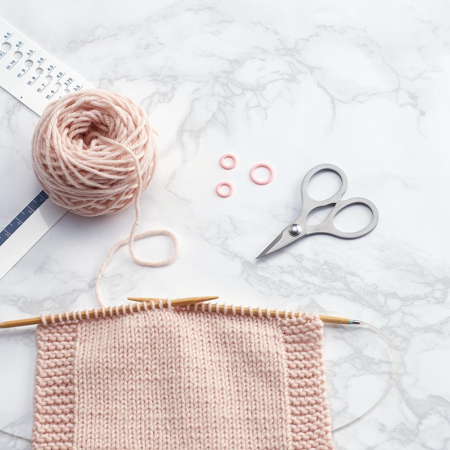 a knitting project in progress, with a pink knitted item, a ball of yarn, and a pair of scissors on a marble surface.