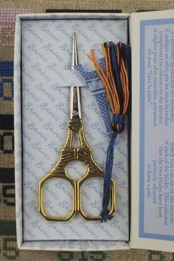 A pair of gold-colored scissors with a blue and orange tassel is displayed in a white box with a blue and white patterned interior.