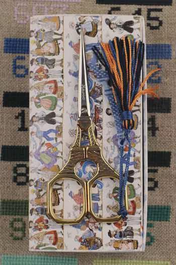 A pair of gold-colored scissors with a blue and orange tassel is placed on a patterned fabric, which appears to be a piece of embroidery or a decorative textile.