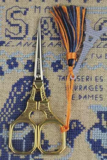 A pair of gold-colored scissors with a tassel attached, placed on a cross-stitch pattern background.