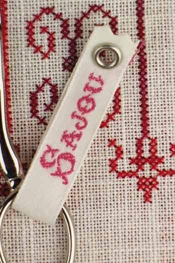 A white keychain with a red embroidered name tag attached to it, resting on a white fabric surface with red embroidered patterns.