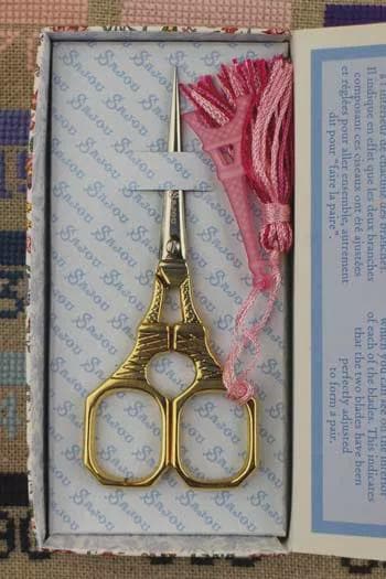 A pair of gold-colored scissors with a pink tassel is displayed in a box with a blue and white patterned background.