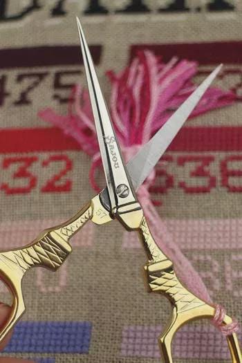 A close-up image of a pair of gold-colored scissors with a pink tassel attached, against a background with a patterned fabric.