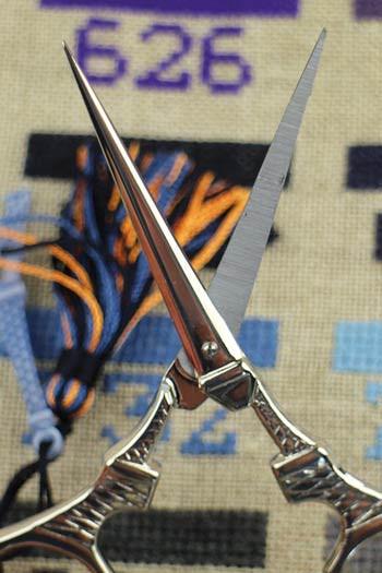 A pair of silver scissors with a colorful tassel is prominently featured in the foreground, with a background of a fabric with a purple and blue pattern.
