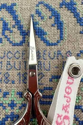 A pair of brown scissors with a white handle is placed on a beige fabric with blue and green embroidered designs.