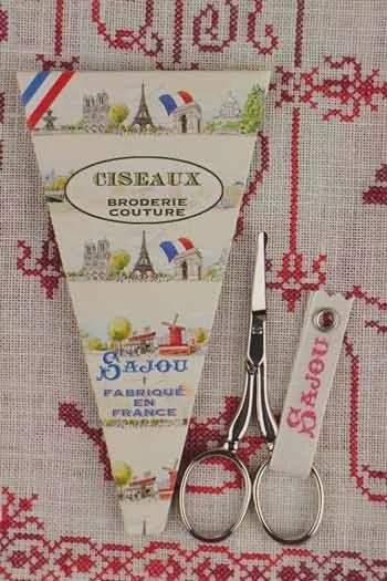 a pair of scissors and a French-themed box, likely a sewing kit, placed on a red and white patterned fabric.