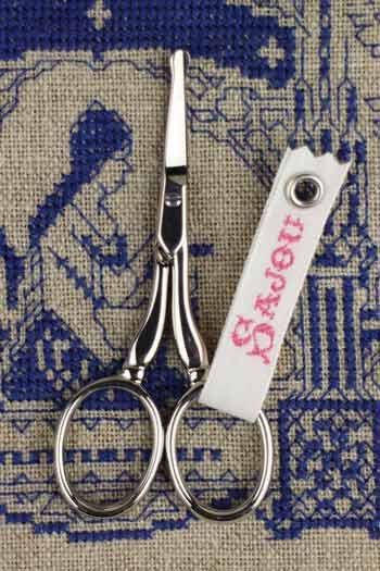 A pair of silver scissors with a white tag attached to them, placed on a blue and white embroidered fabric background.