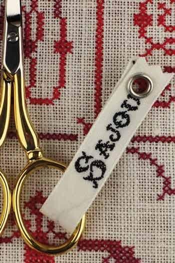 A close-up view of a pair of gold scissors with a white tag attached to them, resting on a red and white embroidered fabric.
