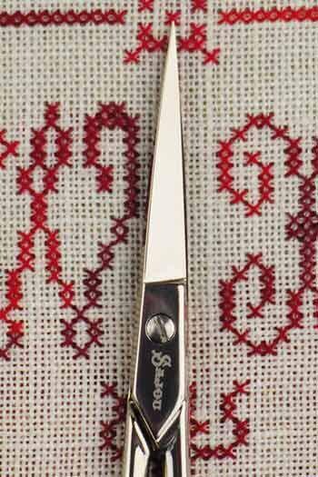 A pair of scissors with a silver handle and a white blade is shown against a background with red and white embroidered patterns.