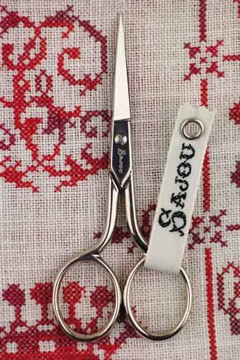 A pair of silver scissors with a white tag attached to them, placed on a red and white embroidered fabric.