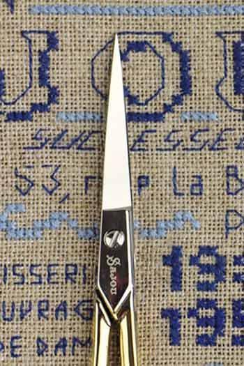 a pair of scissors with a silver handle and a black cross on the blades, placed on a textured fabric with blue and white embroidered patterns.