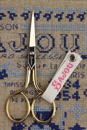 A pair of gold-colored scissors with a white tag attached, resting on a cross-stitch pattern with blue and white colors.