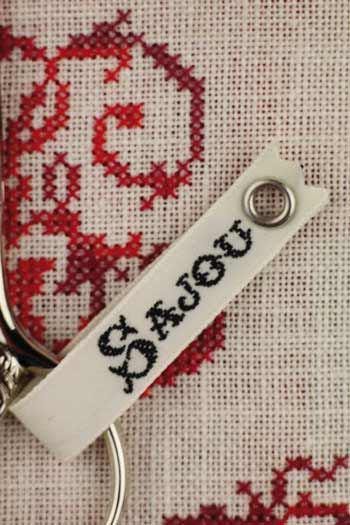 a close-up view of a white keychain with a metal loop, attached to a beige fabric with a red and white embroidered design.
