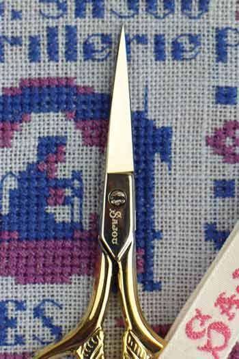A close-up image of a pair of gold-colored scissors resting on a cross-stitch pattern.