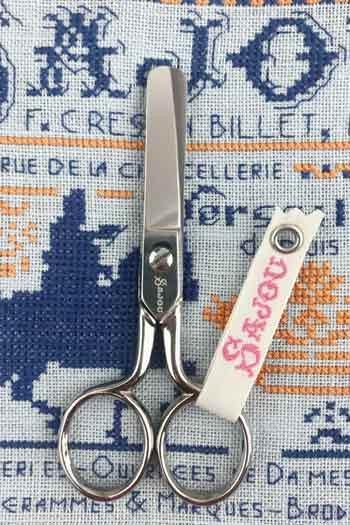 A pair of scissors with a white tag attached to it, placed on a fabric with a cross-stitch pattern.