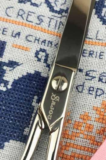 A close-up view of a pair of scissors with a metal handle and blades, resting on a fabric with a cross-stitch pattern.