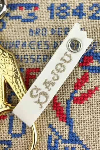 a close-up view of a white keychain with a gold-colored metal clasp, resting on a beige fabric with a blue and red embroidered pattern.
