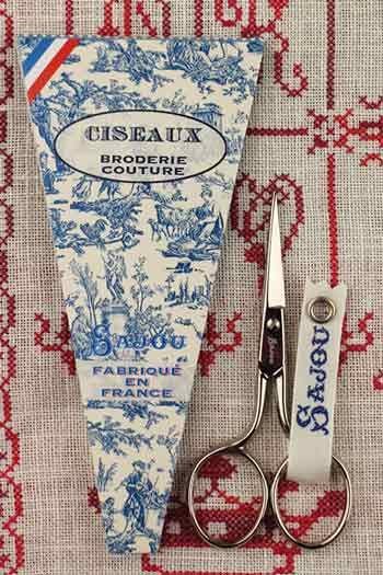a pair of scissors and a package of fabric scissors, both with a French-inspired design, placed on a red and white patterned fabric background.