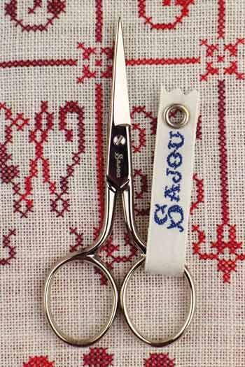 A pair of scissors with a white tag attached to them, placed on a red and white embroidered fabric background.