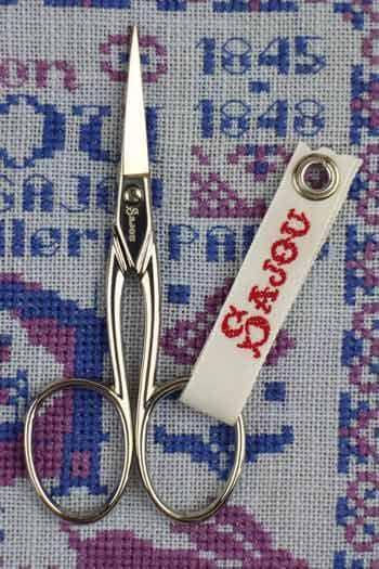 A pair of scissors with a white tag attached to them, placed on a cross-stitch pattern background.