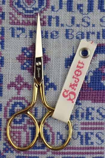 A pair of gold-colored scissors with a white tag displaying the word "Dover" is placed on a blue and purple embroidered background.
