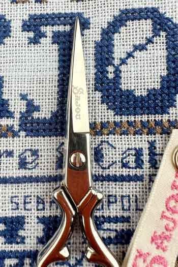 A pair of scissors with a silver handle and a red blade is placed on a blue and white embroidered fabric.