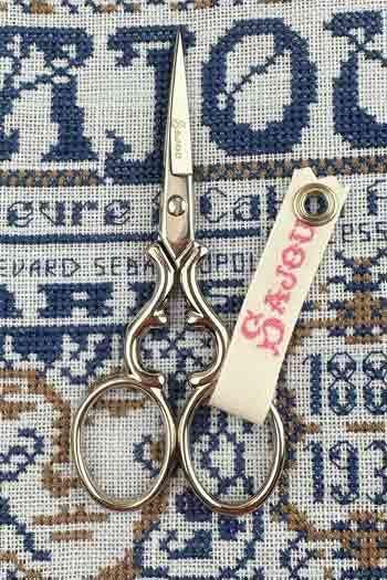 A pair of scissors with a white tag attached to it, resting on a cross-stitch pattern background.