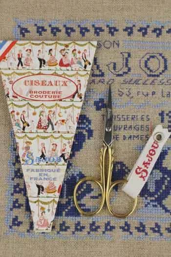 a pair of gold scissors, a cross-stitch kit, and a small box of fabric, all placed on a beige background with a blue and white pattern.
