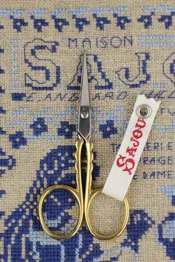 A pair of gold-handled scissors with a white tag displaying the word "SAJOU" is placed on a blue and white cross-stitch pattern.