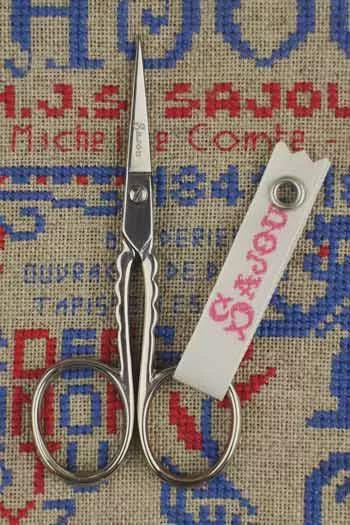A pair of silver scissors with a white tag attached to them, placed on a beige fabric with a blue and red pattern.