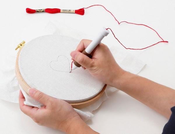 A person's hands are holding a hoop with a heart-shaped design on it, and they are using a marker to draw a heart on the fabric.
