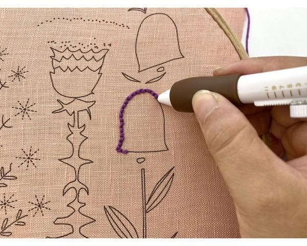 A hand is using a purple needle to embroider on a fabric with various floral and leaf designs.