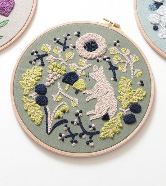 a circular embroidery piece with a floral design, including a pink flower, green leaves, and other decorative elements.